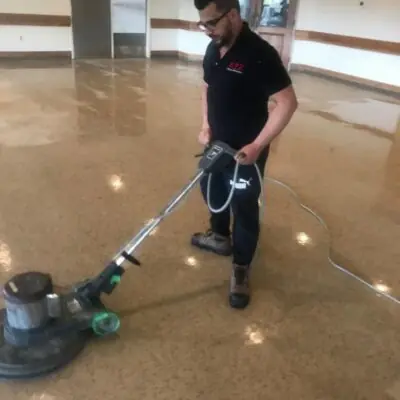 Man in work attire operating a floor polishing machine in a large indoor space.