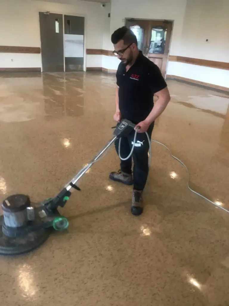 Man in work attire operating a floor polishing machine in a large indoor space.